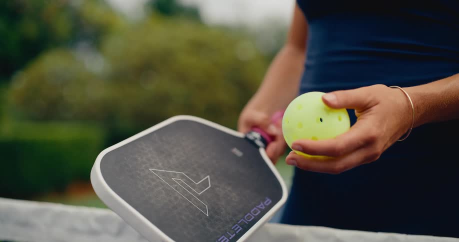 A woman bounces a yellow pickleball on her paddle in extreme slow motion on an outdoor court