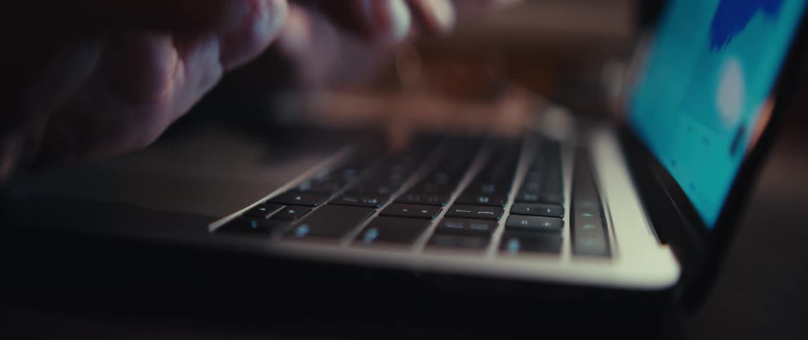 A slow rack focus close-up shot of hands typing on a laptop keyboard in a dimly lit environment