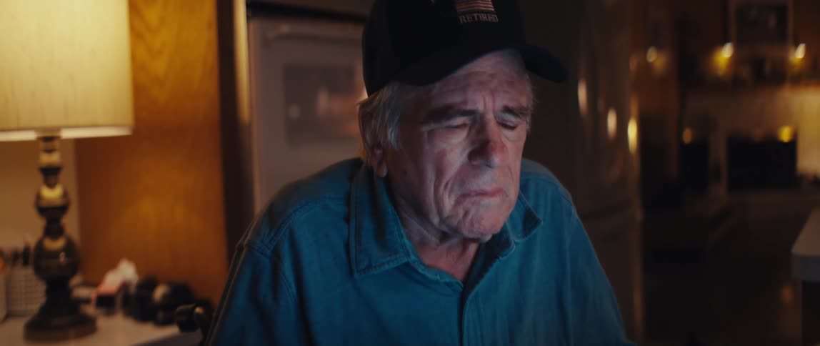 A close-up on the sad face of an elderly retired firefighter tilts down as he sits in front of a laptop at night