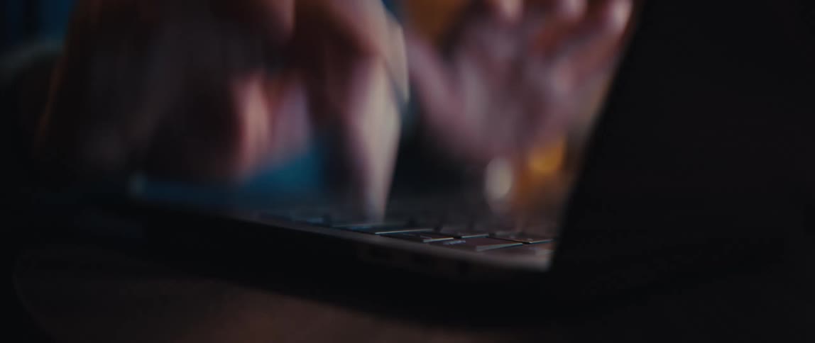 A close-up, out-of-focus shot reveals a person's hands typing on a laptop keyboard in a dimly lit room with warm bokeh lights