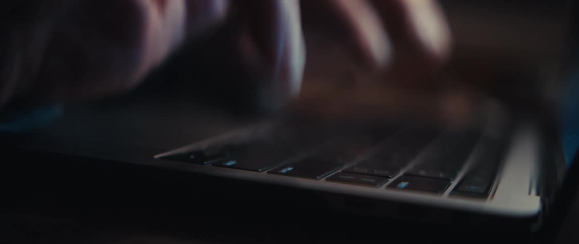 A dramatic rack focus reveals a close-up of hands typing on a laptop keyboard in low light