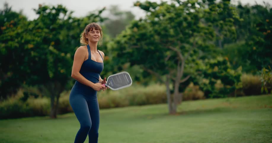 An athletic woman in blue sportswear smiles after hitting a powerful overhead pickleball shot in slow motion on an outdoor court