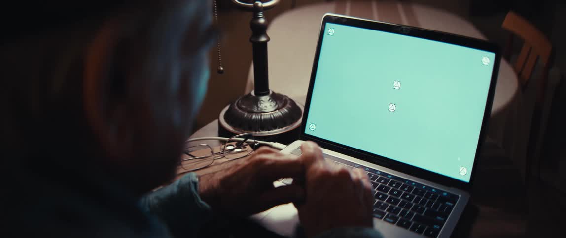 Over-the-shoulder shot of an elderly man looking at a laptop with a green screen in a dark room