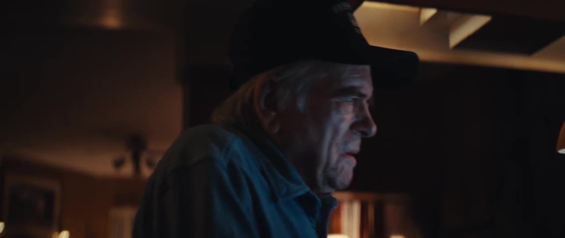 A close-up of a serious elderly man in a cap watching a screen in a dimly lit room at night