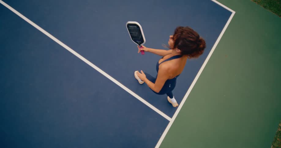 From above, a woman in athletic wear hits a pickleball in slow motion as a futuristic data graph appears on her paddle