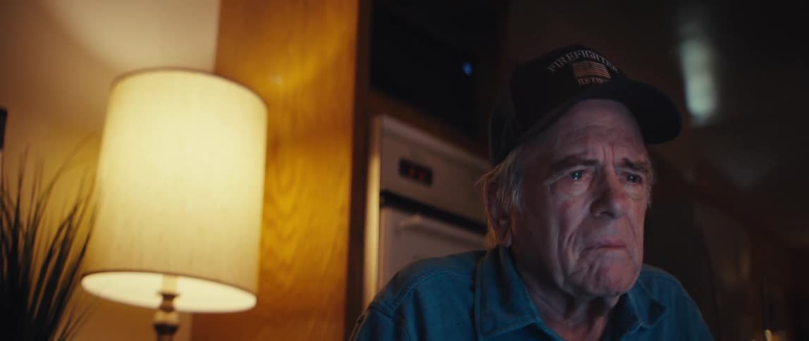 An elderly retired firefighter gazes intently at a screen, his face filled with somber emotion
