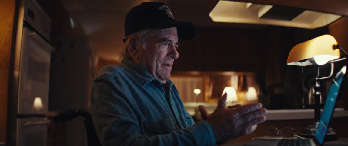 An elderly man in a wheelchair smiles as he opens his laptop to video chat with family in his cozy home at night