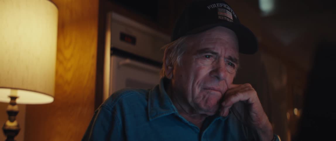 A pensive elderly retired firefighter sits alone in a dimly lit room watching something off-screen with a somber expression