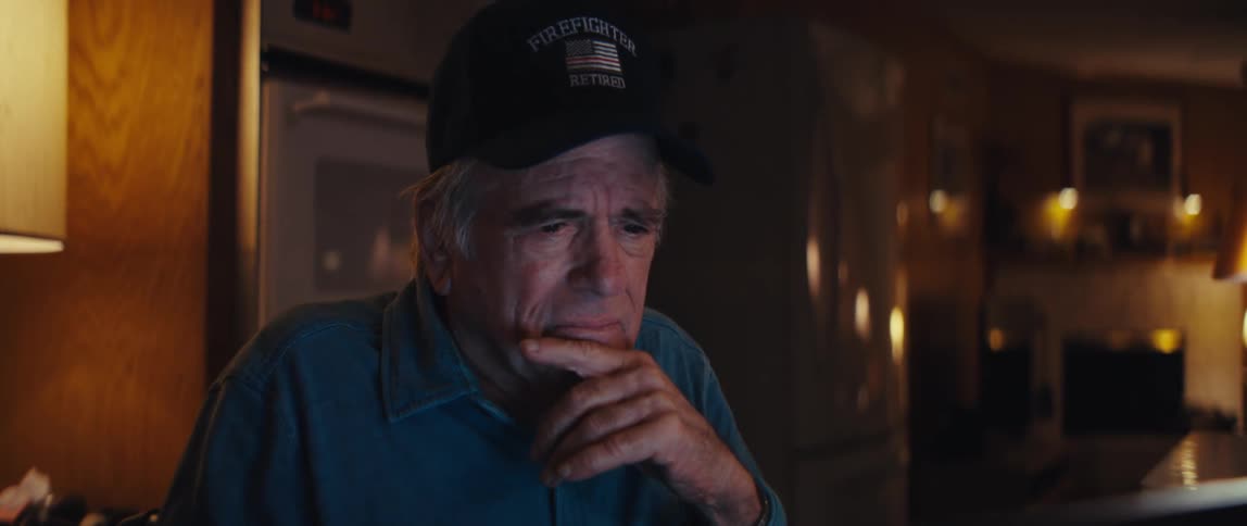 A pensive elderly man in a 'Retired Firefighter' hat stares at his laptop screen late at night in a dimly lit room