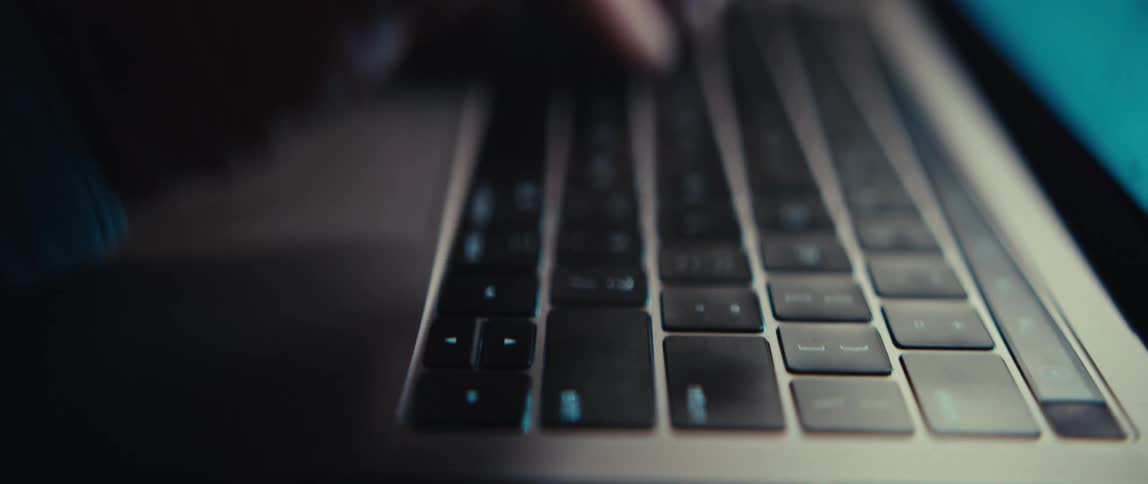 An extreme close-up of hands typing on a laptop keyboard in a dark room, with the camera panning down to the shift and return keys
