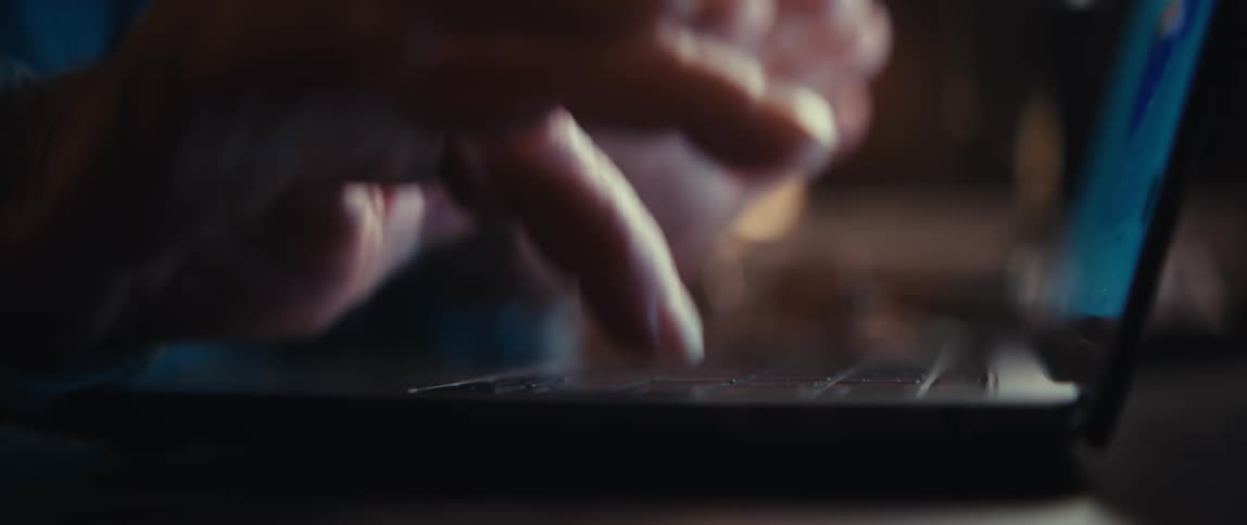 A rack focus close-up shot shows hands typing on a laptop keyboard in a dimly lit room at night