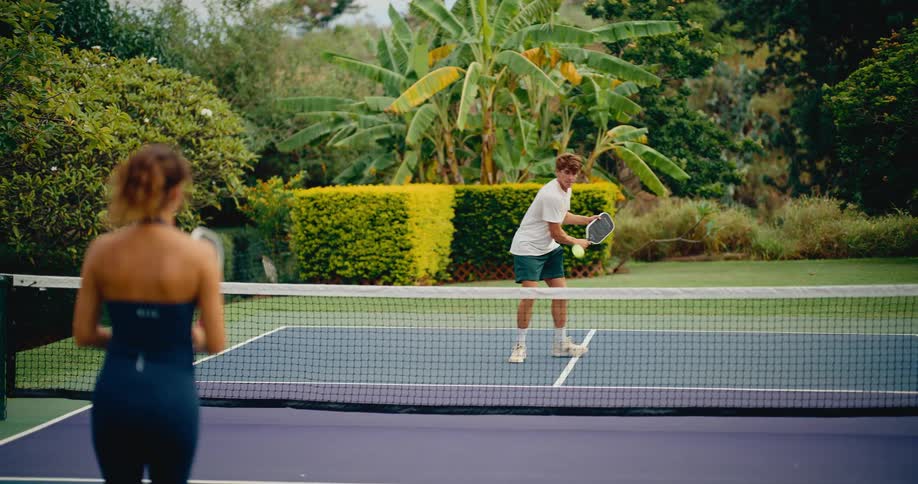 A young man and woman play a game of pickleball on an outdoor court surrounded by lush greenery in slow motion