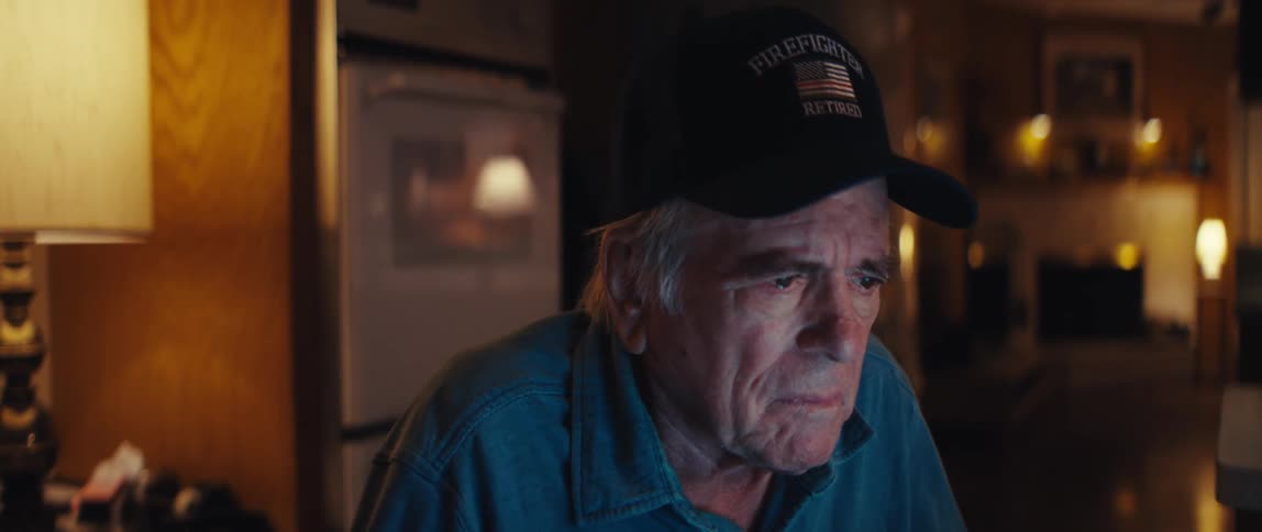 An elderly retired firefighter with a concerned expression focuses intently on a screen in his home at night