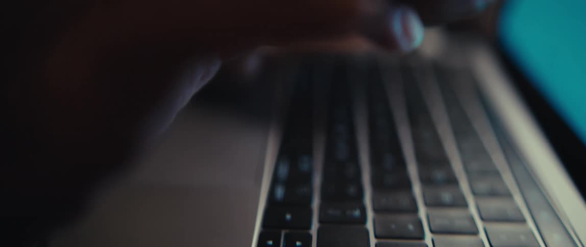 A shallow focus, close-up shot shows the wrinkled hands of an elderly person typing on a laptop keyboard in a dimly lit room
