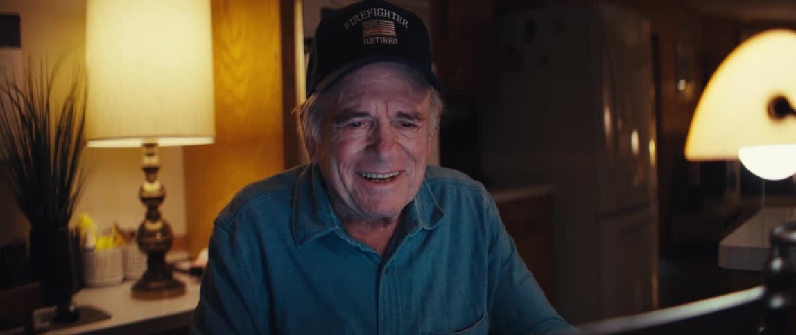 A happy elderly man, a retired firefighter, smiles warmly while using his laptop for a video call in his cozy home at night