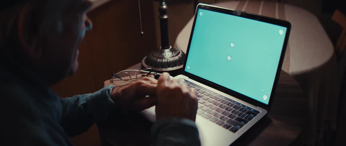 Over-the-shoulder shot of an elderly man typing on a laptop with a green chroma key screen in a dimly lit room
