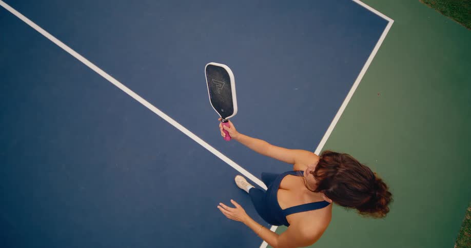 A top-down slow-motion view of a woman serving a pickleball on a blue and green court