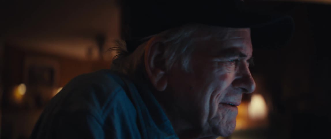 An elderly man works on his laptop at home at night, his face lit by the screen