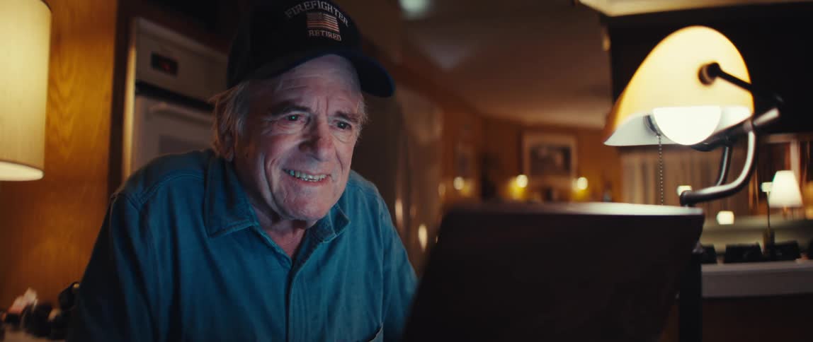An elderly man wearing a retired firefighter hat smiles warmly while looking at his laptop screen in a cozy home at night