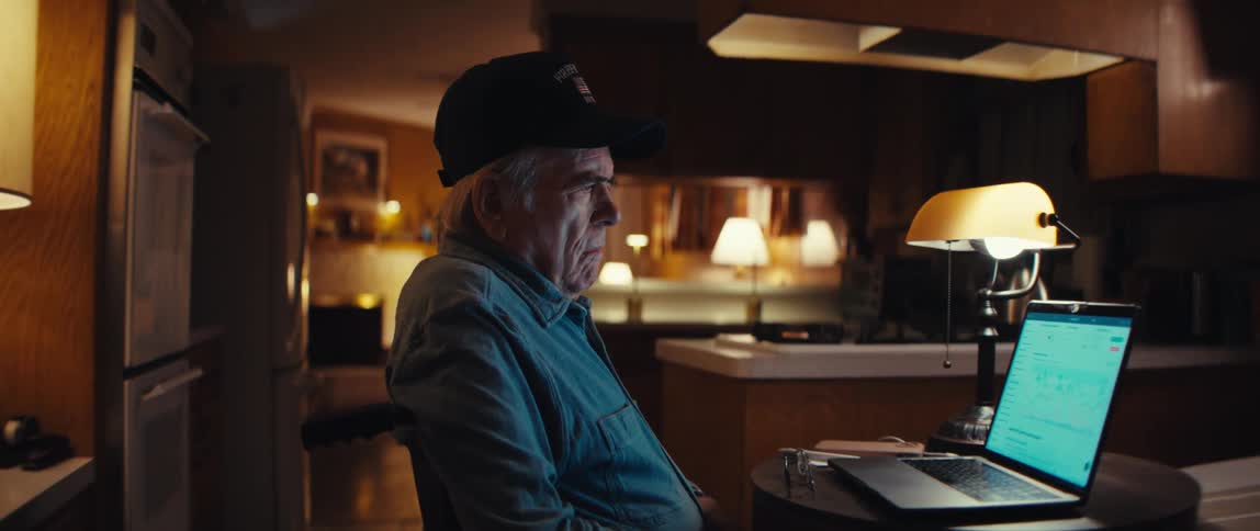 A senior man in a wheelchair sits alone in his dimly lit home, intently focused on his laptop screen