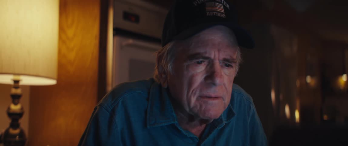 A close-up of a concerned elderly man wearing a retired firefighter hat as he watches something on a screen in a dark room