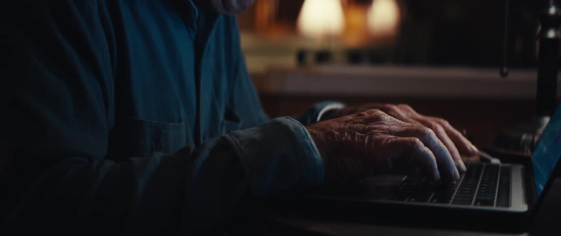 A slow-motion close-up captures the wrinkled hands of a senior citizen typing on a laptop keyboard at night