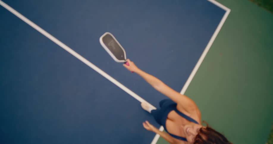 A female athlete serves a pickleball in slow motion, captured from a dynamic top-down perspective on a colorful court