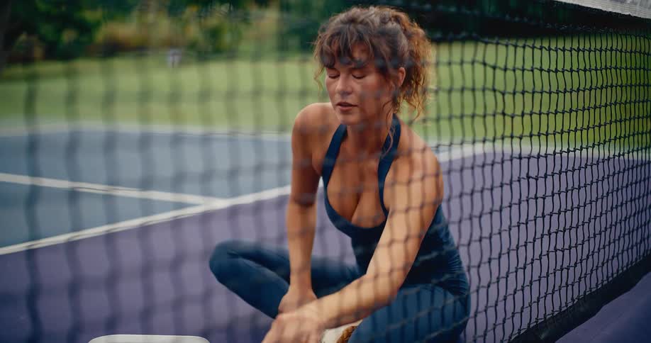 View through the net of a thoughtful woman in athletic wear resting on an outdoor tennis court