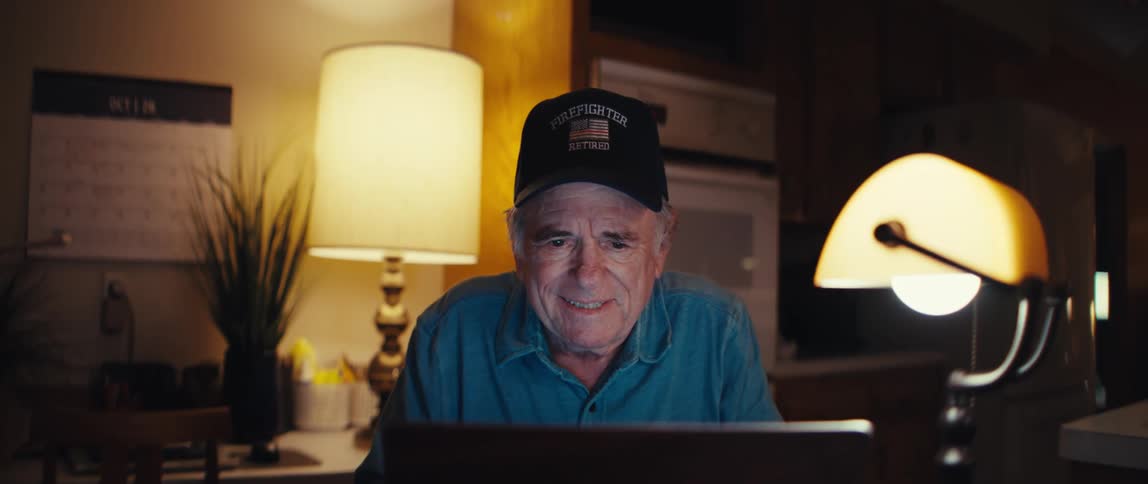 A smiling elderly man, a retired firefighter, uses his laptop at the kitchen table late at night