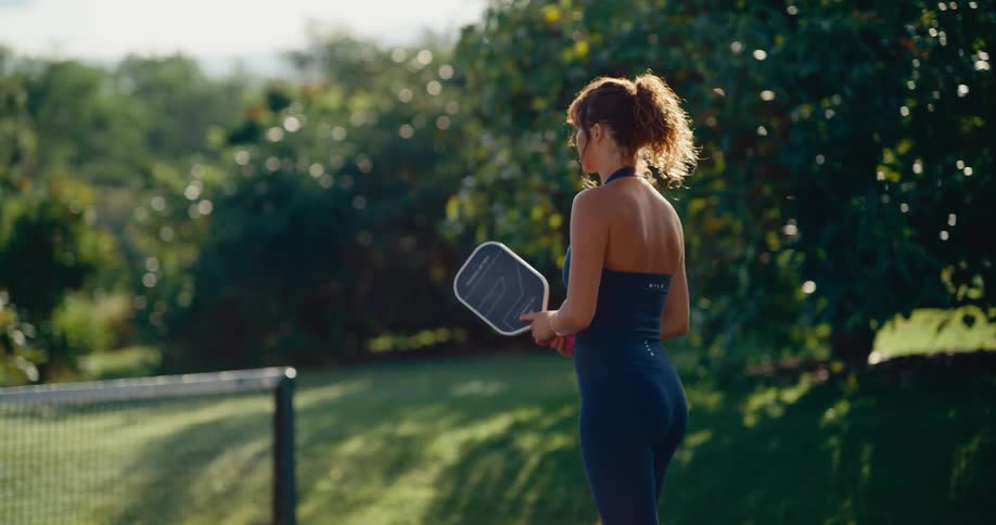 A woman hits a pickleball in slow motion, seen from behind on a sunny outdoor court