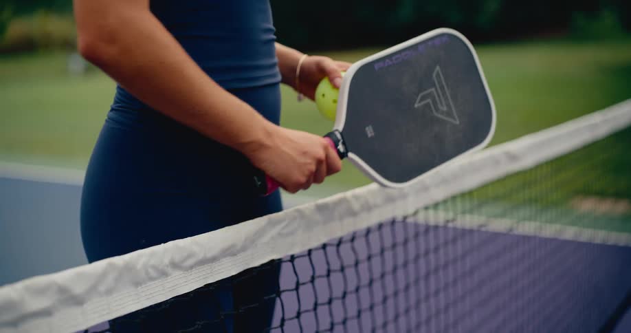 A close-up shot of a female athlete holding a pickleball paddle and ball while standing next to the net on an outdoor court