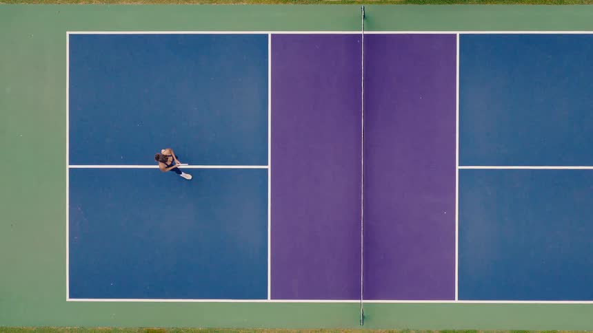 A slow-motion overhead drone shot captures a lone woman gracefully practicing pickleball on a vibrant blue and purple court