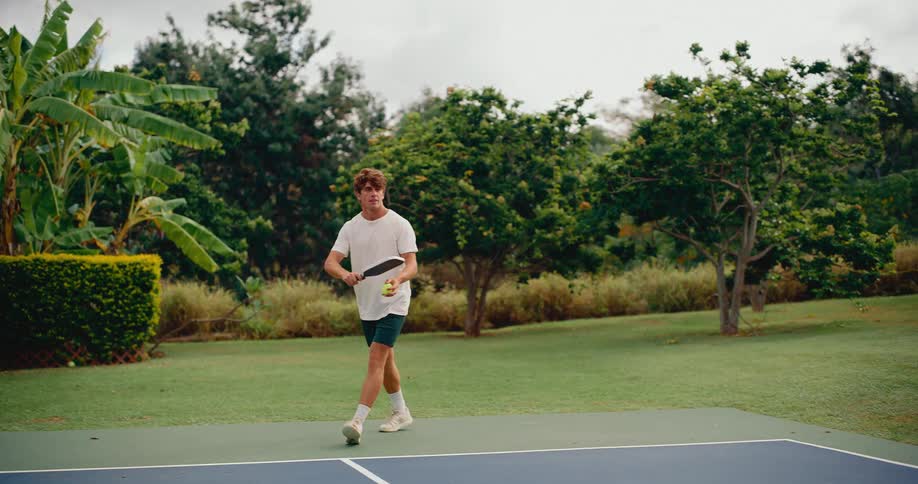A young man serves a pickleball in slow motion on a beautiful outdoor court surrounded by lush greenery