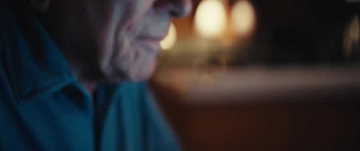 An elderly man's focused face, lit by a laptop screen, tilts down to reveal his wrinkled hands typing on the keyboard at night