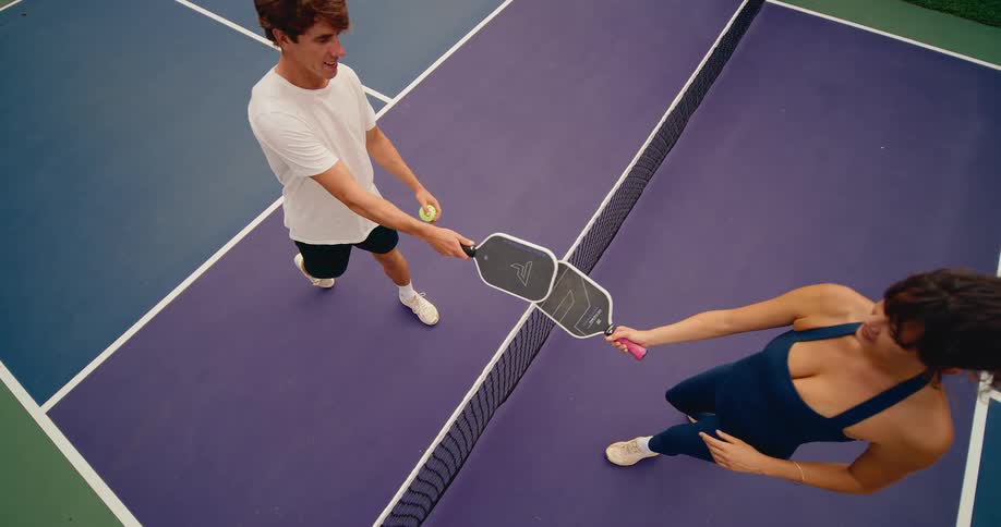 A man and woman demonstrate good sportsmanship by touching their pickleball paddles over the net in a friendly top-down slow-motion shot