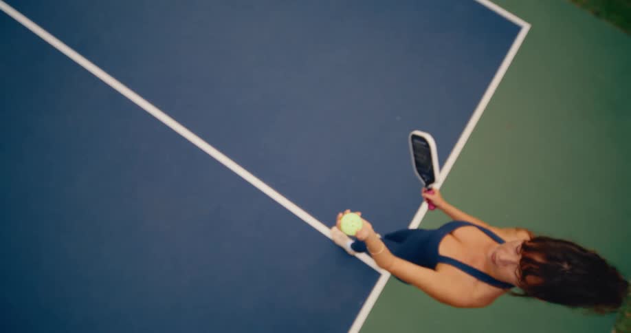 Slow motion top-down view of a woman on a pickleball court tossing the ball up to serve