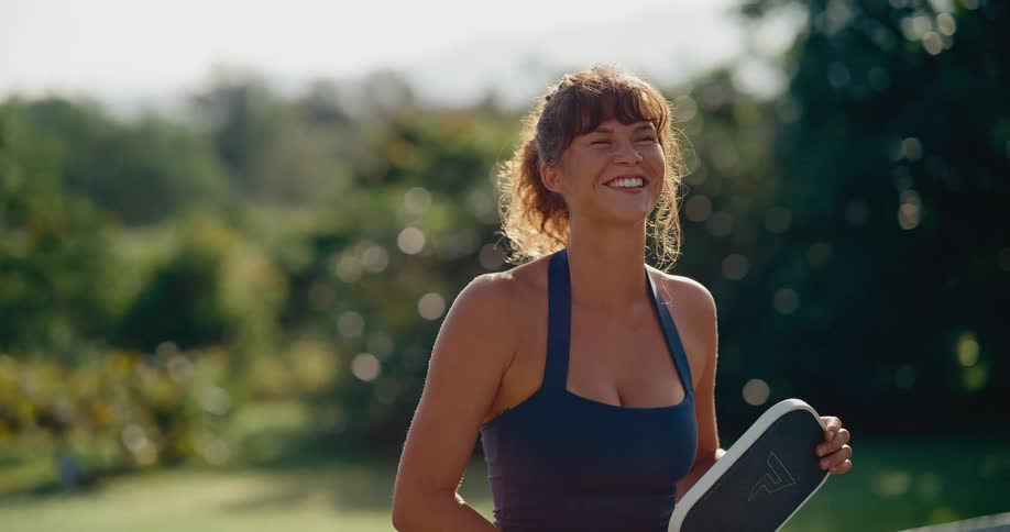 A joyful and athletic woman smiles brightly in the sun before playfully preparing for a game of pickleball in slow motion