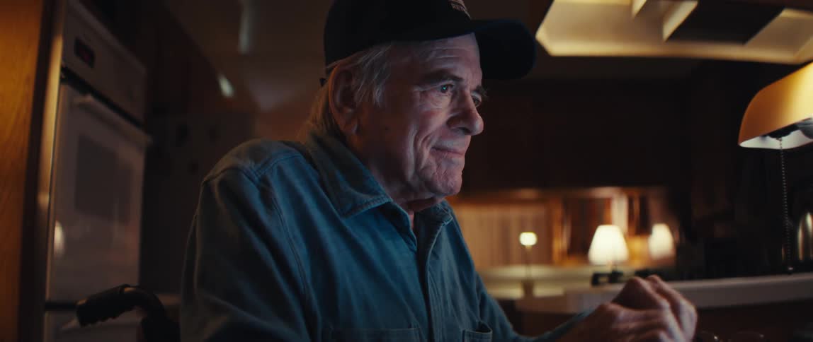 A smiling elderly man in a wheelchair opens his laptop at a table, his face lighting up as he connects with someone online