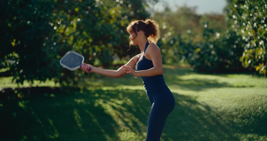 An athletic woman smiles as she hits a pickleball with a paddle in slow motion on a beautiful sunny day
