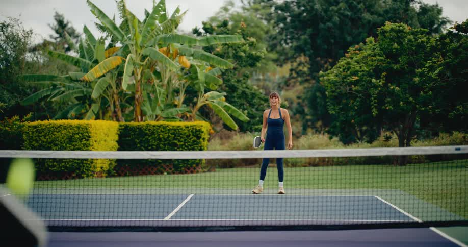 A woman hits a forehand shot in slow motion while playing pickleball with a man on a beautiful private court surrounded by lush greenery