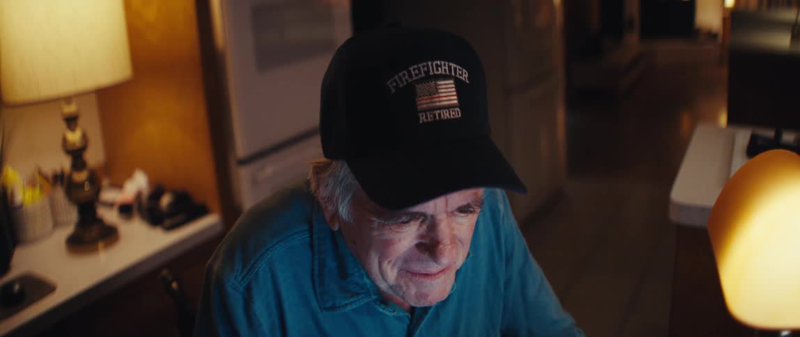 A happy retired firefighter in a wheelchair smiles while typing on a laptop computer at his desk in a dimly lit room