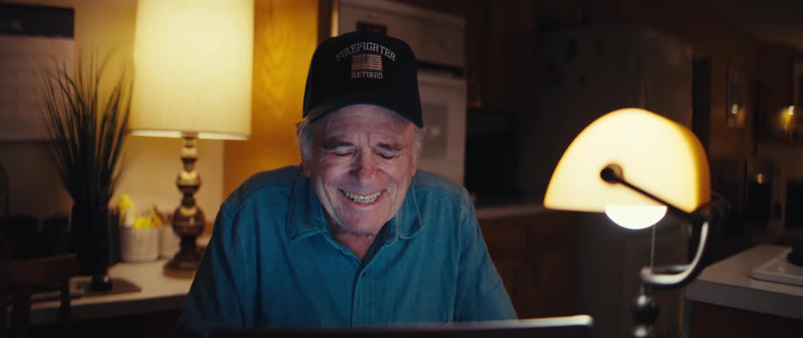 An elderly retired firefighter's joyful expression turns to sadness while he types on his laptop at night