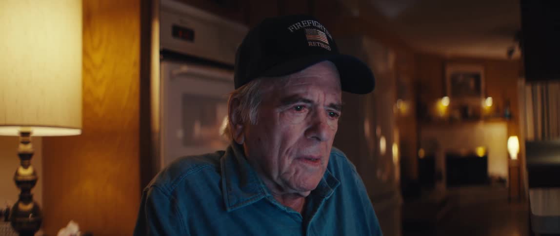 An emotional close-up shows a retired firefighter with a sad expression watching something in his home