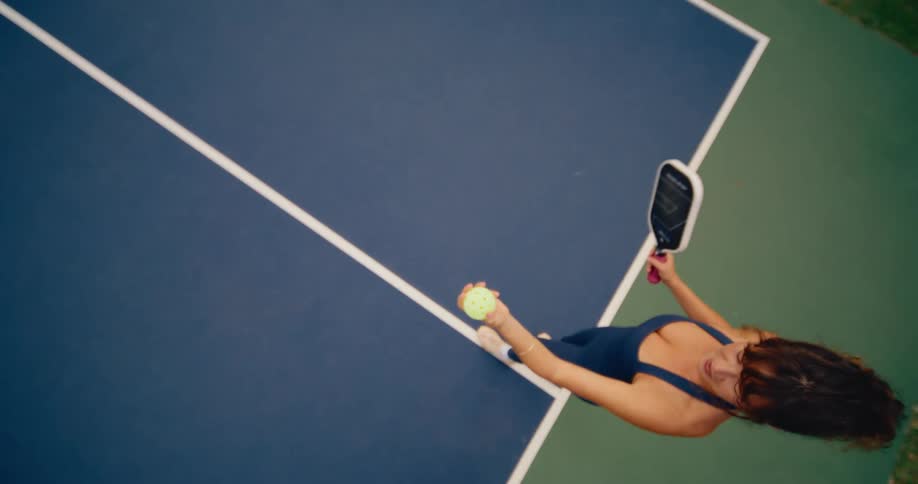 Slow motion top-down view of a happy woman tossing a pickleball into the air on a pickleball court