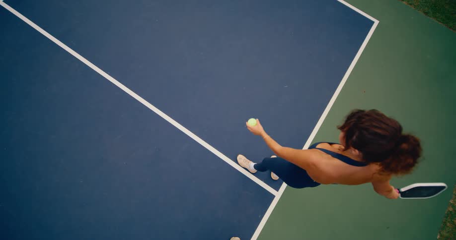 A top-down slow-motion view of a woman in blue athletic wear serving a pickleball on a blue and green court