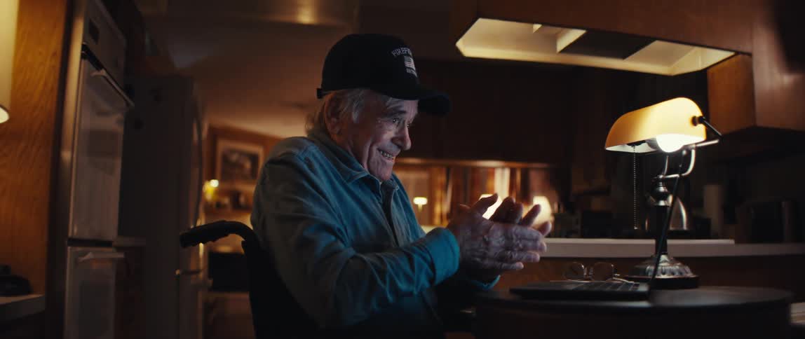 An elderly veteran in a wheelchair smiles as he opens his laptop to video chat with someone in his home at night