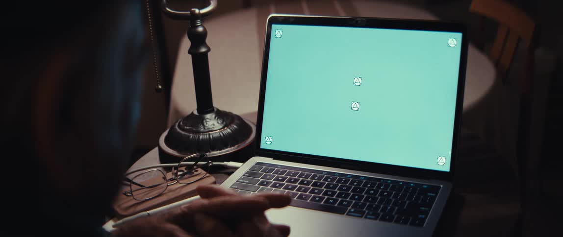 An elderly person watches a green screen laptop at a desk in a dimly lit room