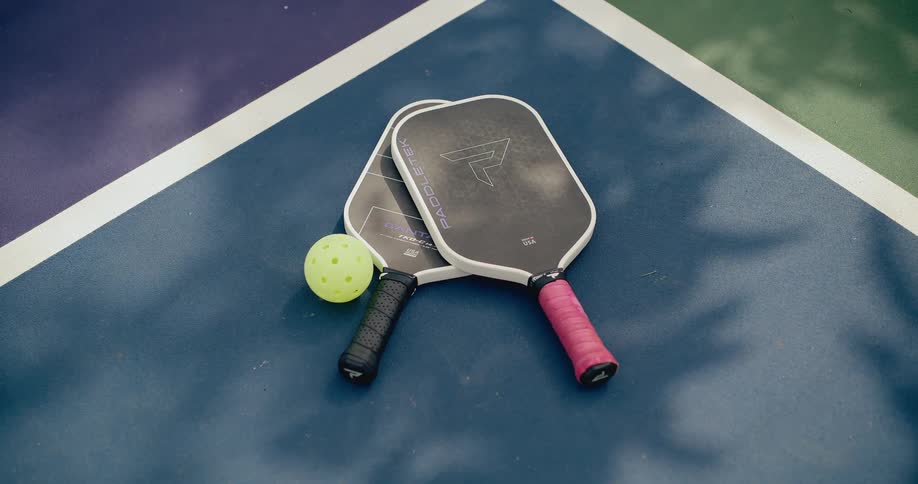 Two pickleball paddles and a ball rest on a blue court as tree shadows dance in the sunlight