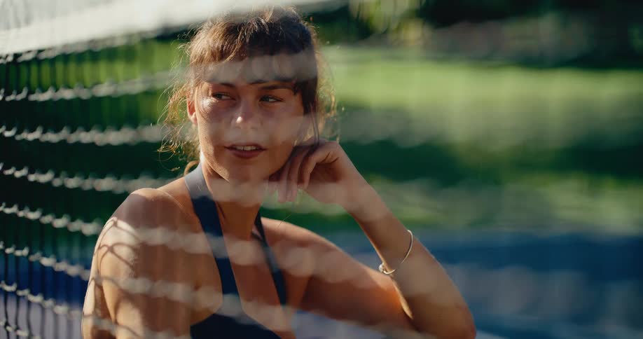 A thoughtful young woman with curly hair relaxes by a tennis net on a sunny day