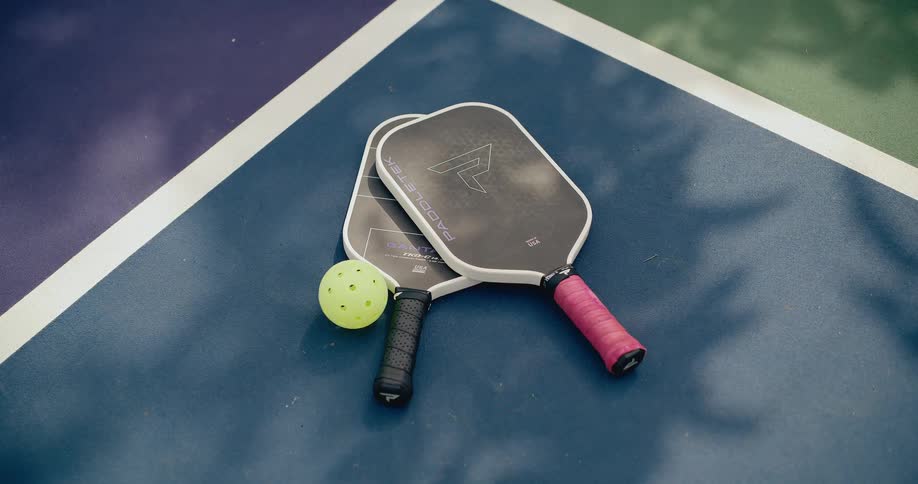 Two pickleball paddles and a bright yellow ball rest on a blue court as shadows from trees dance across them